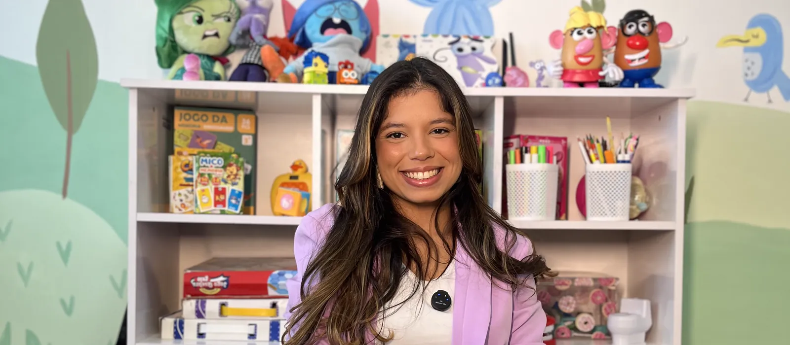 Jessica Costa sorrindo no consultório do Vertice Mall, Recreio dos Bandeirantes: estante com pelúcias dos personagens Divertida Mente, jogos pedagógicos e mural infantil ao fundo