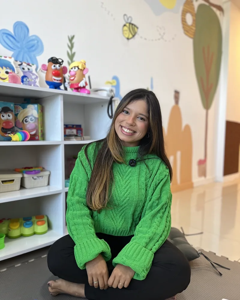 Jessica Costa, psicóloga de crianças e adolescentes, sentada na sala lúdica do consultório com sorriso aberto, blusa verde, brinquedos coloridos e mural de abelhinha ao fundo