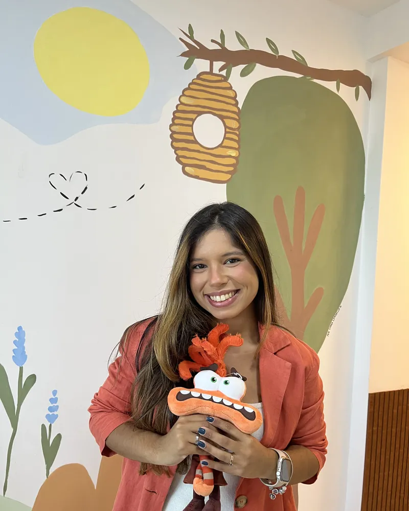 Jessica Costa Psi, psicóloga de crianças e adolescentes, sorrindo em seu consultório com brinquedo terapêutico em mãos e mural infantil ao fundo